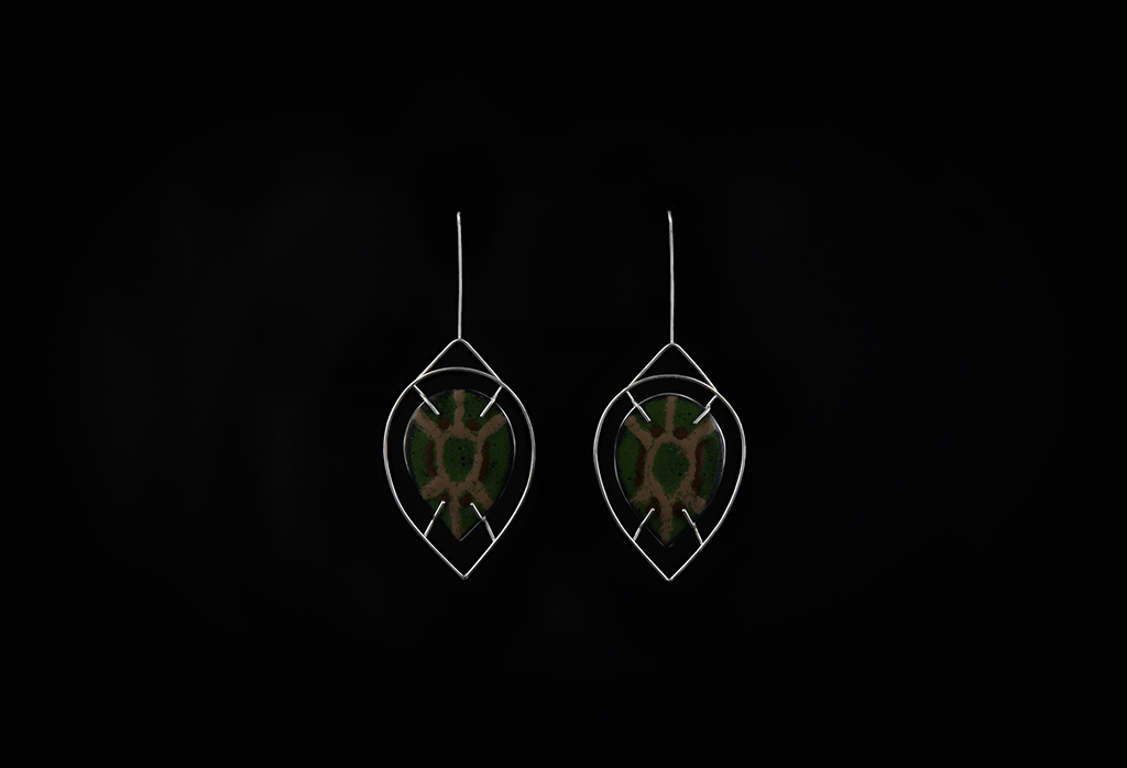 Enamel earrings. In the center is a teardrop shape that resembles a turtle shell. It is a green background with tan and brown lines. This enamel piece is framed by a silver wire frame and looks like it is floating.